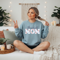 8. Baseball Mom Light blue crewneck sweatshirt with embroidered "Baseball Mom" design featuring script lettering above bold varsity-style letters with authentic baseball stitch detailing in white and red, displayed on a female model seated on a white couch.
