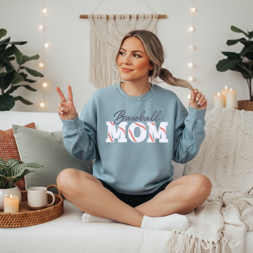 8. Baseball Mom Light blue crewneck sweatshirt with embroidered "Baseball Mom" design featuring script lettering above bold varsity-style letters with authentic baseball stitch detailing in white and red, displayed on a female model seated on a white couch.
