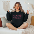 Senior Class of 2026 Light blue crewneck sweatshirt with embroidered "Senior Class of 2026" text featuring a decorative bow accent in pink, displayed on a female model seated on a white couch.