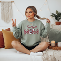 Football Mom Gray crewneck sweatshirt with embroidered "Football Mom" design featuring script lettering above bold varsity-style letters with a decorative patterned football in maroon and white, displayed on a female model seated on a white couch.