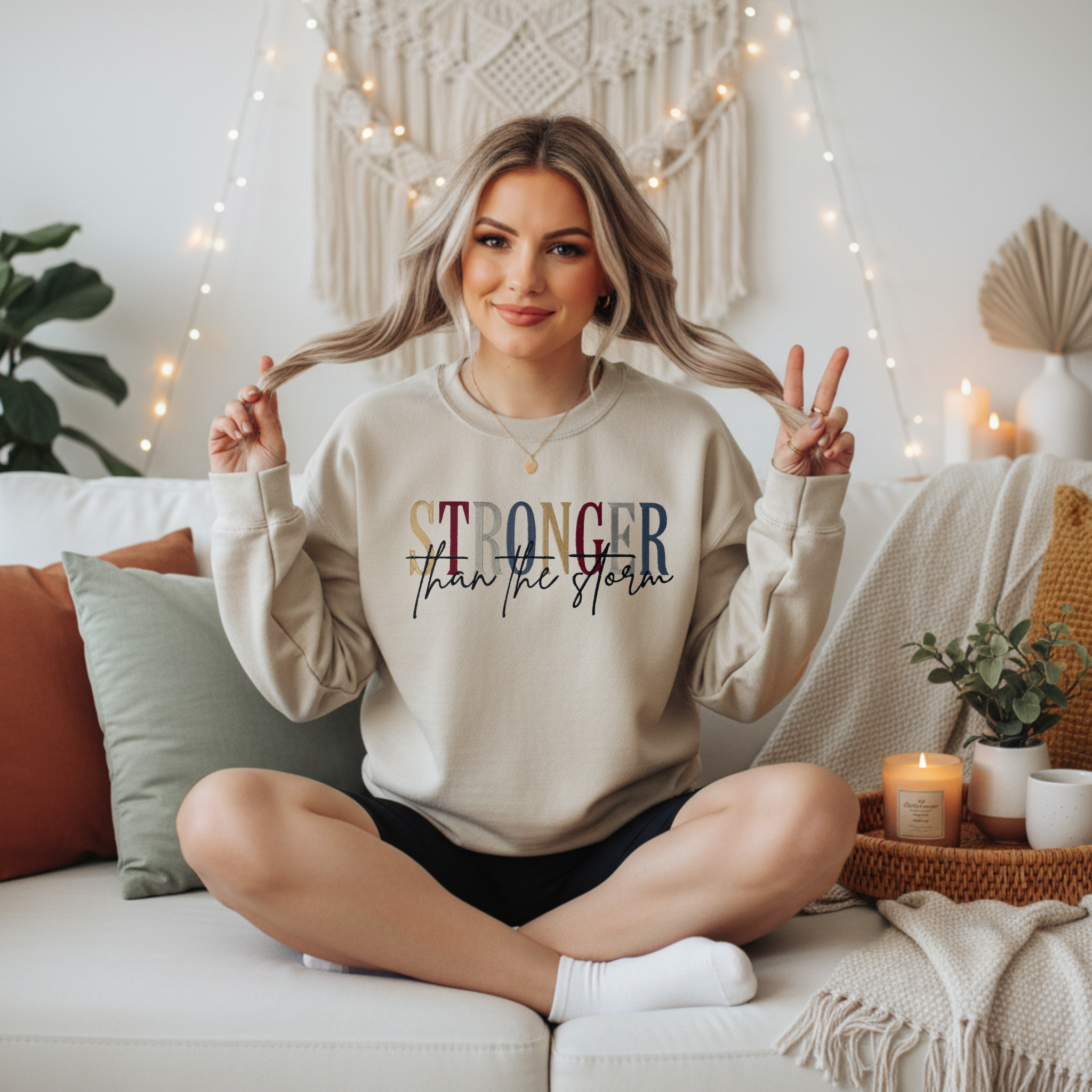 Stronger Than the Storm crewneck sweatshirt with multicolor embroidered "Stronger Than the Storm" motivational text in bold and script lettering, displayed on a female model seated on a white couch.