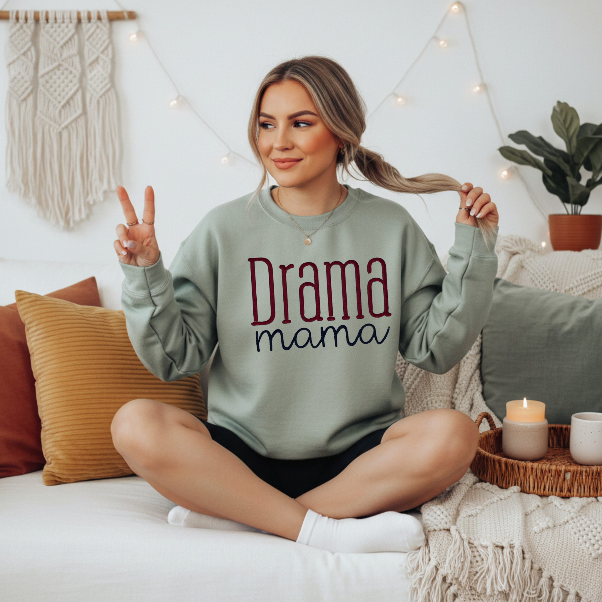 Sand crewneck sweatshirt with embroidered "Drama Mama" design in mixed serif and script lettering in maroon and navy, displayed on a female model seated on a white couch.