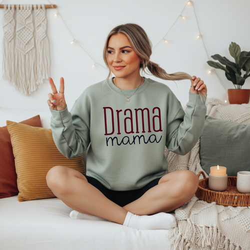 Sand crewneck sweatshirt with embroidered "Drama Mama" design in mixed serif and script lettering in maroon and navy, displayed on a female model seated on a white couch.